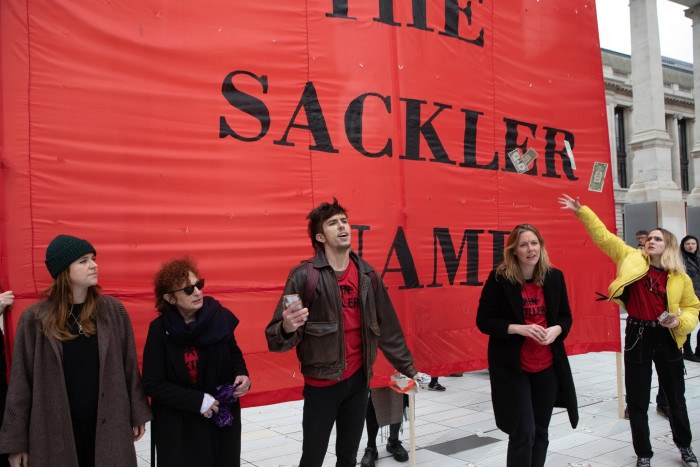 People stand outside a building, in front of a large red sign on which we can see part of the slogan: ‘abandon the Sackler name’