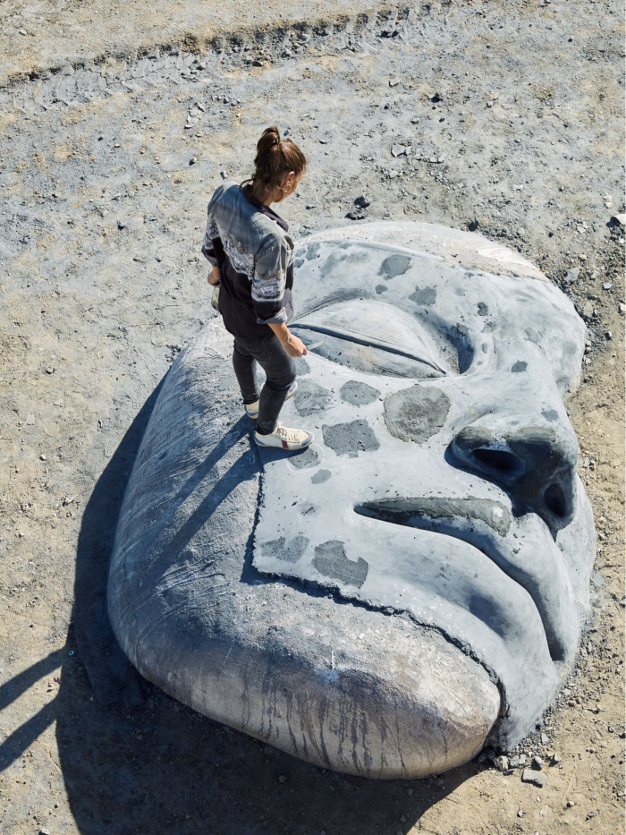 A woman stands on a gigantic clay face lying on the ground