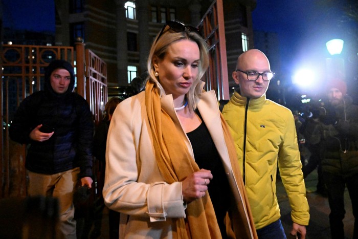 A sombre-looking Marina Ovsyannikova leaves court in darkness, as cameras flash. A man in yellow is with her