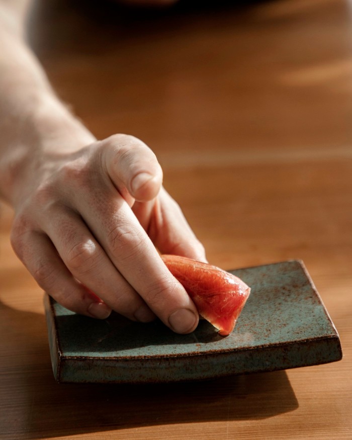 Chef Mads Battefeld’s fingers holding a piece of chutoro nigiri on a small square dish