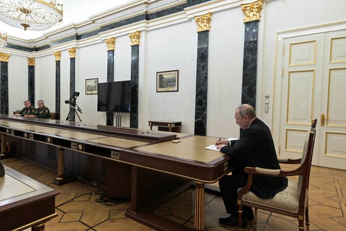 Seated at the near end of a very long table, Putin presides over a meeting with two men in military insignia seated at the far end of the table