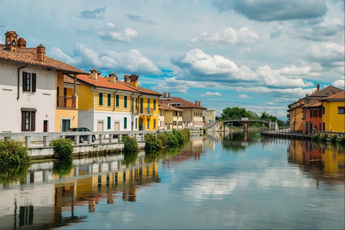 The Naviglio Grande canal running through the town of Gaggiano, with a bridge in the distance and yellow, white and red houses on its banks