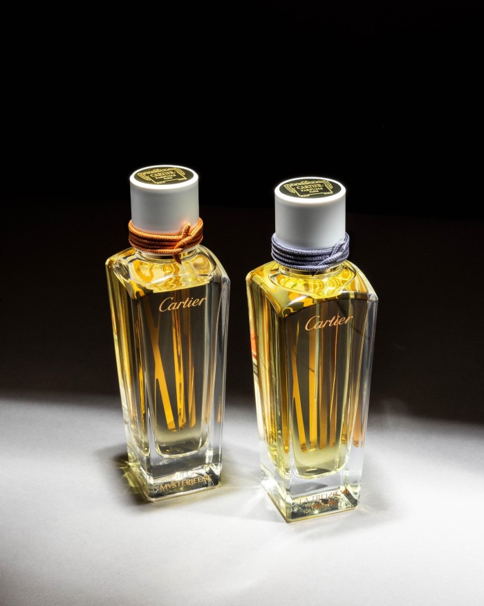 A pair of Cartier perfume bottles, filled with amber liquid, set on a light surface. Each bottle features a distinctive white cap with gold accents and the brand’s emblem, one adorned with an orange band and the other with a purple band, presented against a dark backdrop