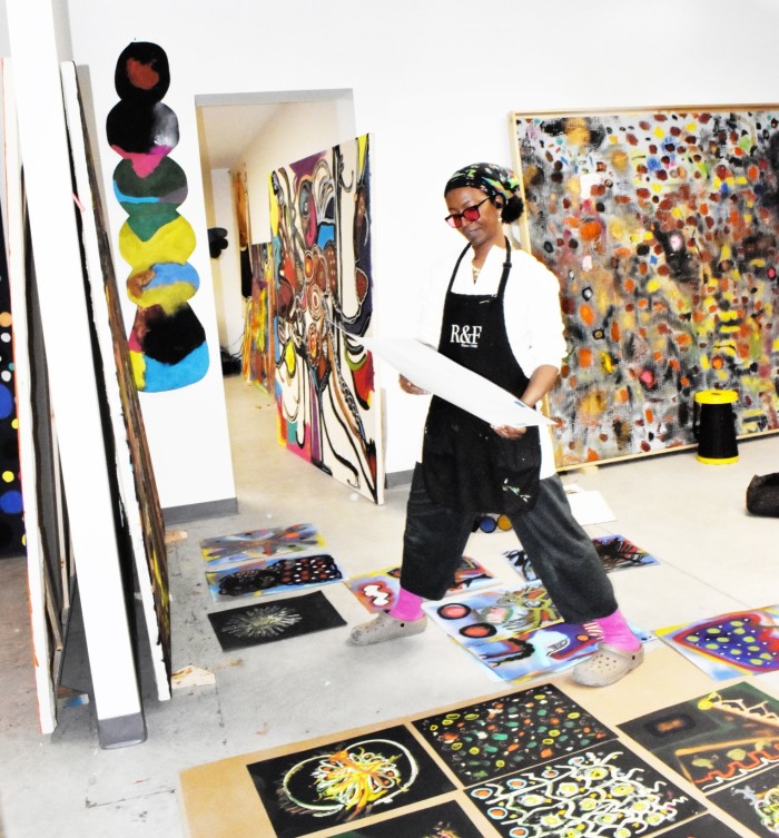 In a photo, a young woman wearing a black apron over a white shirt stands in the middle of her canvases and sketch-filled studio.