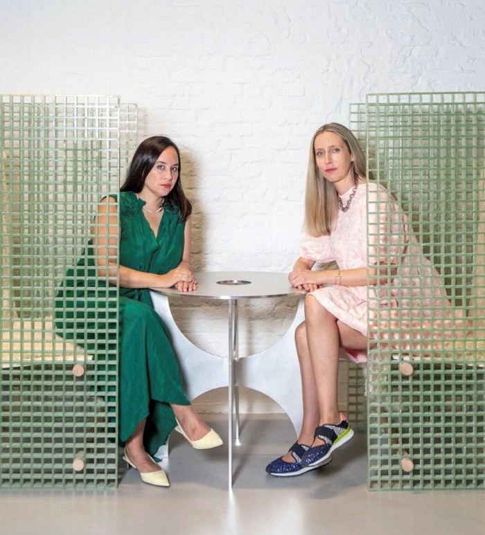 Two women, one dark-haired in a green dress, the other blond in a white dress, sit at a table looking at the camera