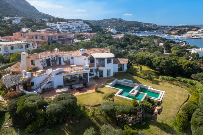A large whitewashed villa with a geometric swimming pool in the garden overlooking a harbour town with many other hillside villas