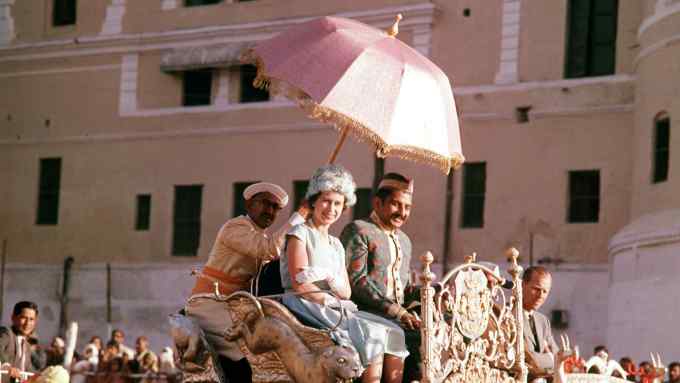 Queen Elizabeth II sits next to a man on an elaborately carved podium that is carried through the streets of India on the back of an elephant. Another man sits behind her, holding a sunshade over her