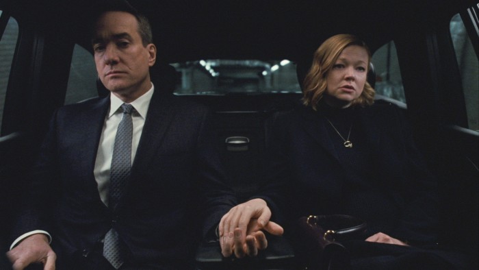 A man and a woman both sharply dressed, holding hands, seated at the back of a car 