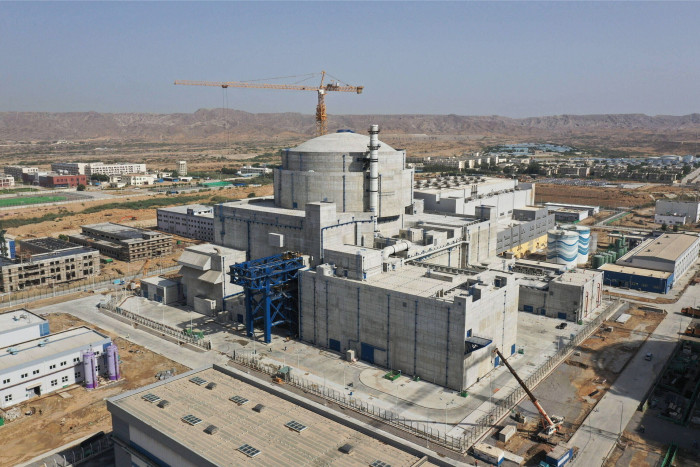 A construction site for a nuclear power plant, showcasing a central domed reactor building surrounded by multiple ancillary buildings and equipment. The scene includes cranes and other machinery, set against a desert-like landscape with mountains in the background