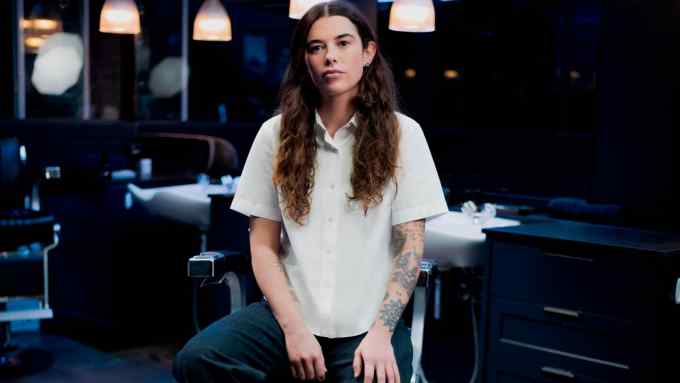 A portrait of a young female barber in a stylish salon setting, featuring dimly lit ambiance, seated confidently with her tattooed arms visible, and wearing a casual white shirt and dark pants
