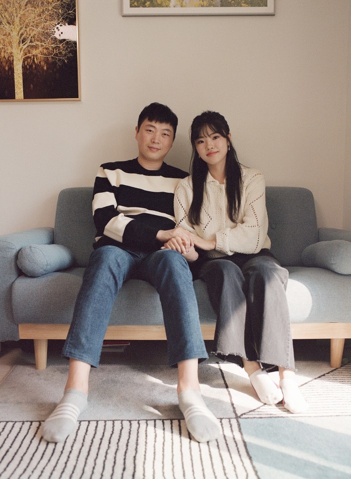 A couple sit close together on a grey sofa in a light filled room, their hands clasped together, looking at the camera and smiling