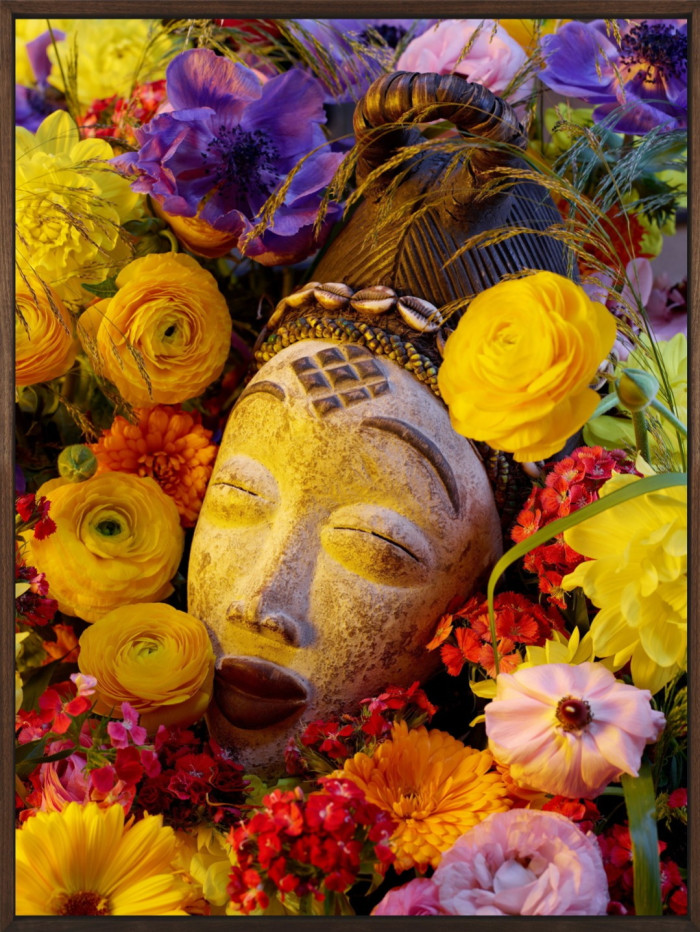 An ancestral wooden mask depicting a human figure with their eyes closed is surrounded by yellow, purple, red and pink flowers
