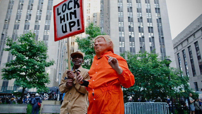 A protester outside the court wearing a costume of prison clothing, a mask of Donald Trump as another protester holds a sign that says ‘lock him up’ 
