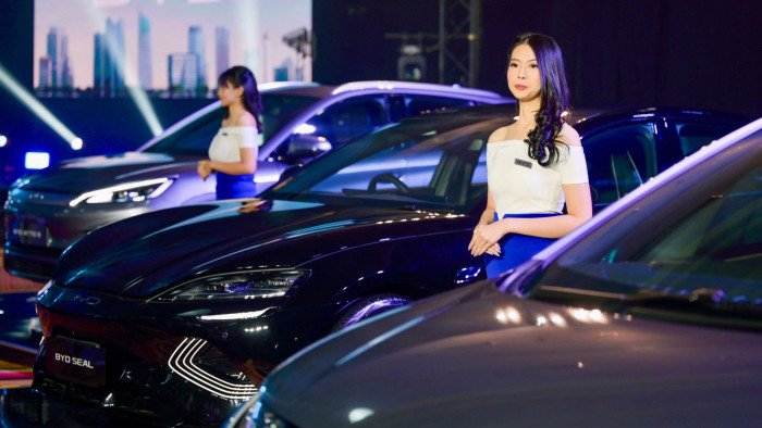 Two women in white tops and blue skirts stand beside Chinese-made electric cars, including a black car labeled ‘BYD SEAL’, at a BYD launch event with a city skyline in the backdrop and dynamic lighting