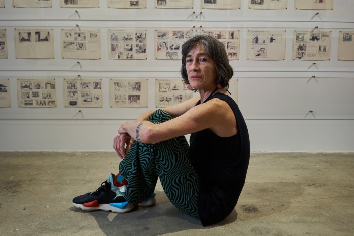 A woman sitting on the ground, legs crossed, in a vest top, with artwork on the wall behind her