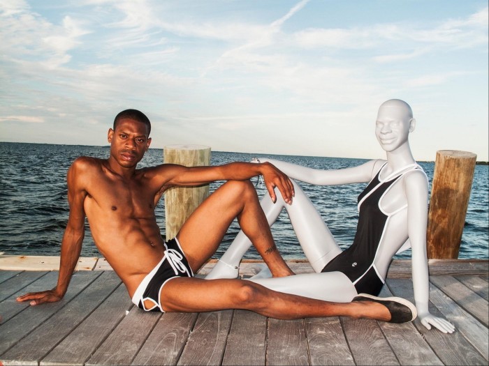 A man in skimpy swimming trunks reclines on a wooden dock next to a mannequin in a swimsuit