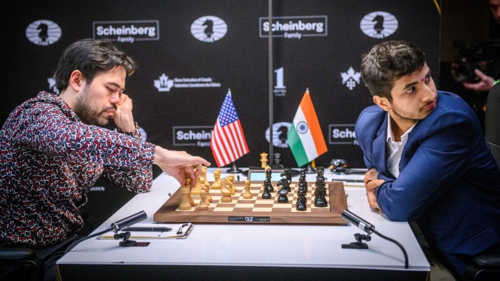 Two young men, both bearded, sit playing at a chessboard. On the table are miniature flags of the US and India