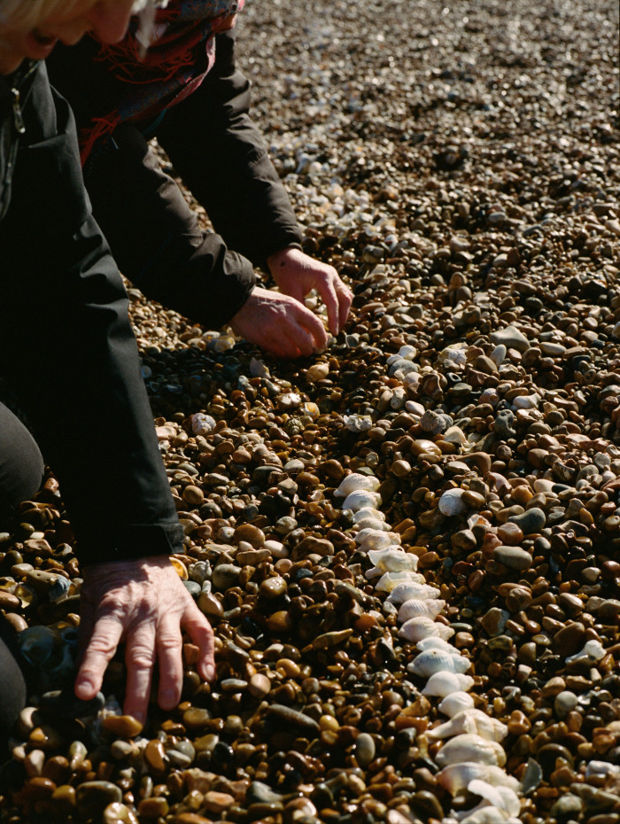 Two people kneel down on the pebbles to arrange shells in a line