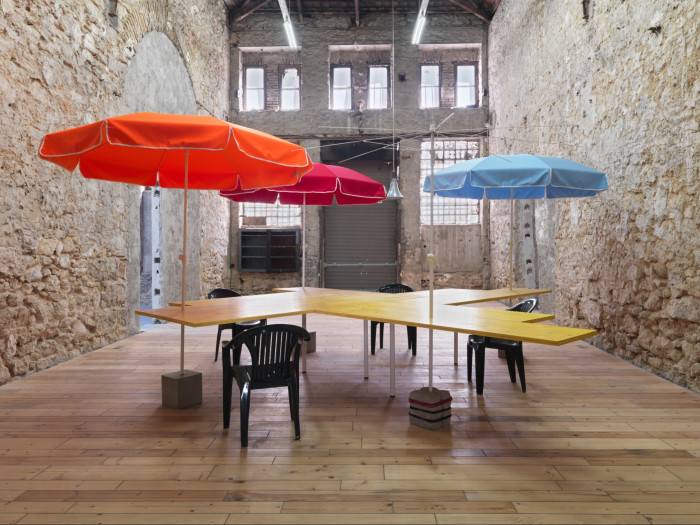 In an artificially lit gallery, a cross-shaped wooden table decorated with bright orange, red and light blue parasols sits in the centre of a room surrounded by four dark plastic chairs.