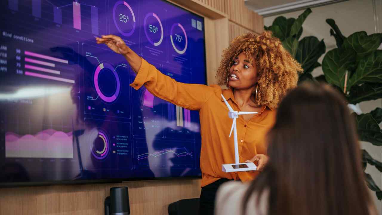 An African American woman is in a boardroom presenting a renewable energy strategy plan and pointing on research displayed on the screen and holding a windmill model in her hands