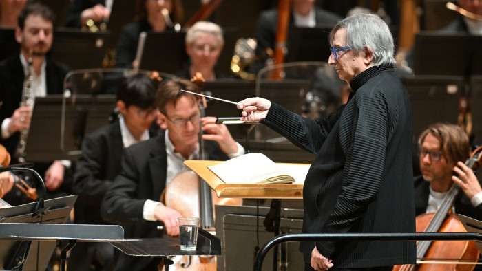 A grey-haired man turns to the side while conducting an orchestra