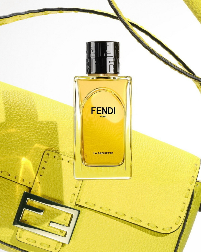 A yellow-coloured rectangular bottle with black shiny lid with a yellow bag with silver clasp in the background