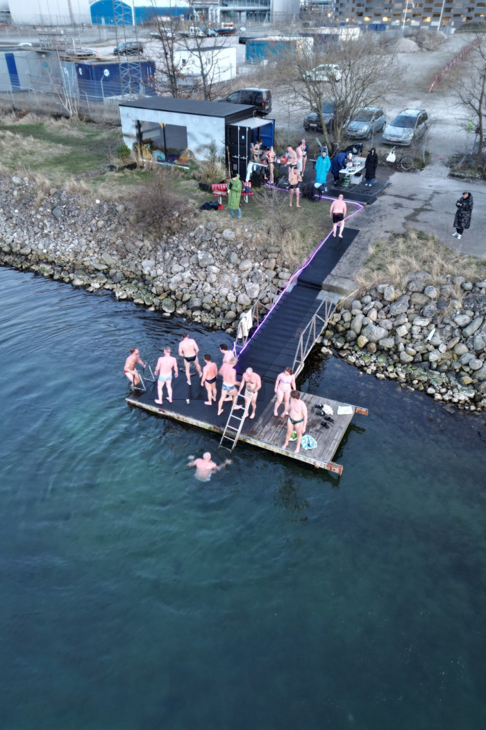 An aerial view of people in swimming costumes on a diving deck near water. In the background is a changing cabin and a car park