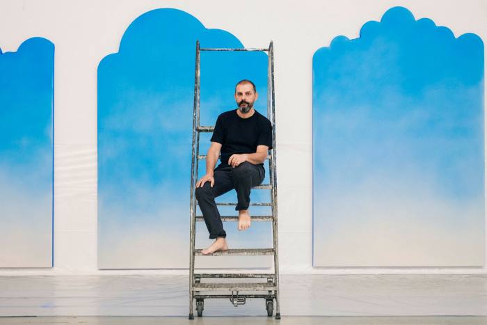 A man sitting on a ladder in front of a series of paintings which look like the sky