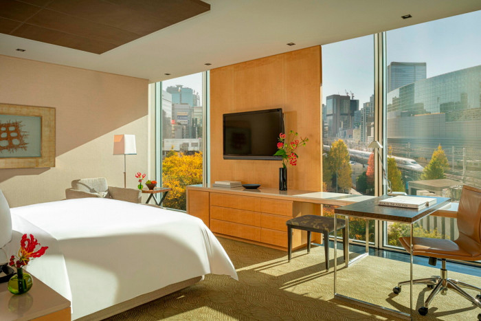 A room at the Four Seasons Marunouchi, with the bed looking towards a blond-wood panel with a TV screen on it, which is flanked by floor-to-ceiling windows looking out towards the city and passing bullet trains