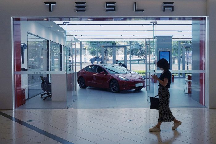 A woman walks past a Tesla showroom while looking at her phone. Inside the showroom, a red Tesla car is prominently displayed with open doors, against a backdrop of large glass windows