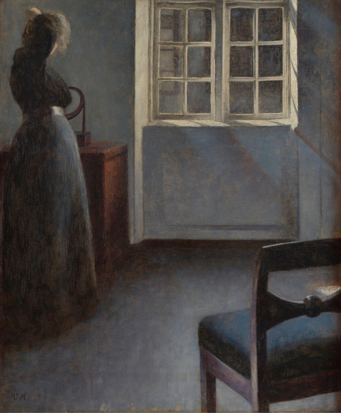 In a night painting, a woman in a dark long dress is seen from behind as she glances out of the window of a room decorated with wooden furniture