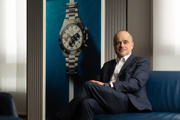 De Clerck sits on a sofa beside a larger-than-life framed picture of a watch