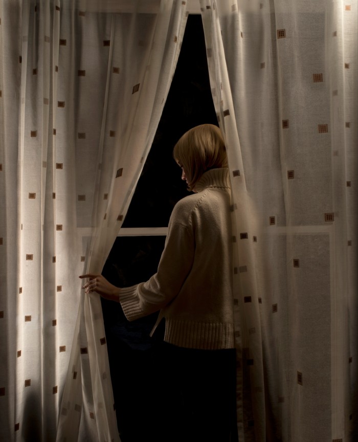A woman stands behind curtains and peers into the darkness outside the window