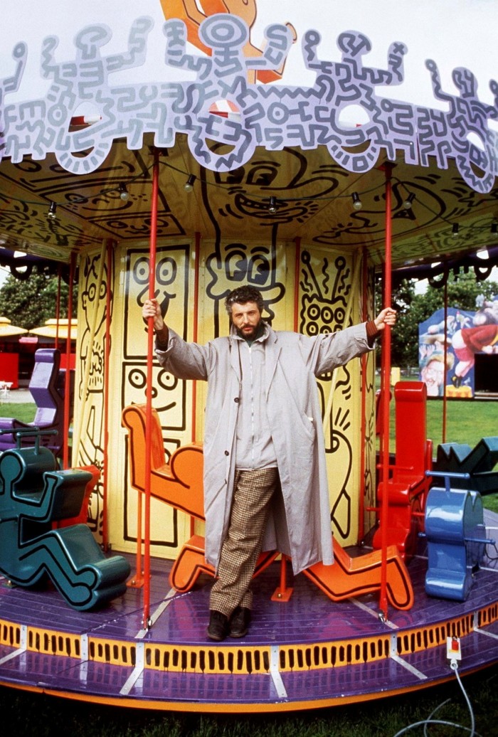 A middle-aged man wearing a long grey coat and chequered trousers stands on a carousel decorated with stylised human figures