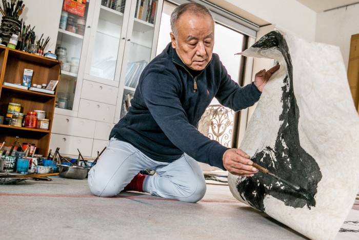 Liu Kuo-Sung kneels in his studio, applying black ink to a textured sheet of paper that he is holding up with one hand 