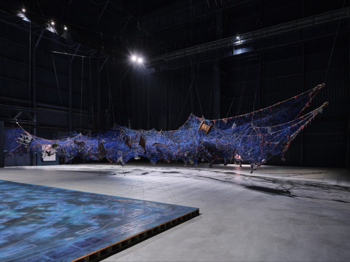 A blue and red textile web comprising wooden, plastic and tin objects floats across a black room which hosts a blue spray-painted platform on its left