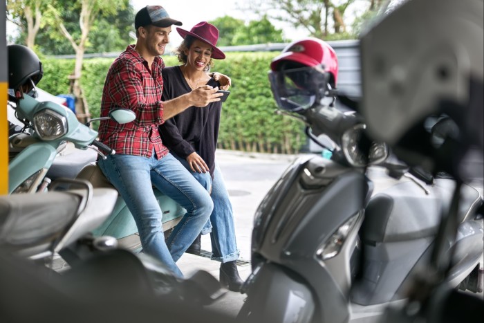 A man and a woman lean against the seat of a motor scooter as they look at a smartphone