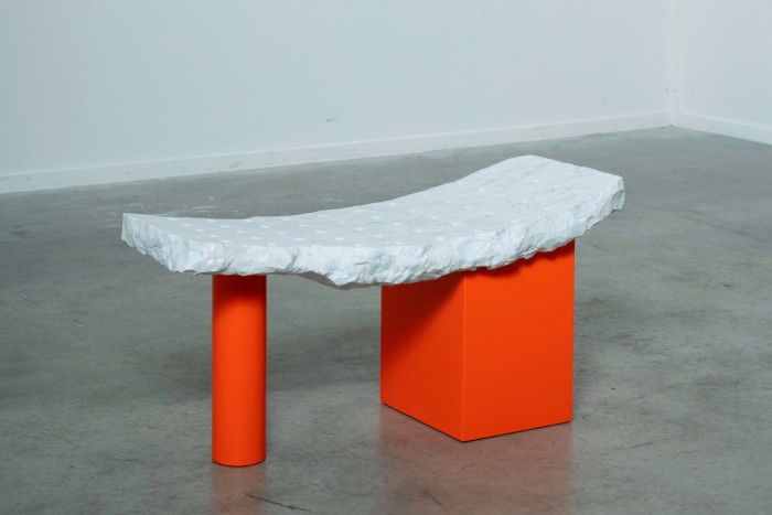 A rough-hewn white bench on orange legs, one a small cylinder, the other a big block