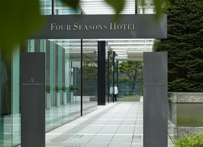 Two grey columns at the entrance to the Four Seasons at Marunouchi, with a doorman in a white coat standing at the far end of a glass wall. In the foreground around the edge are leaves