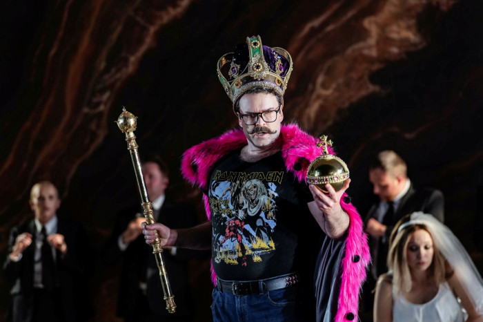 A man with a suspicious moustache, carrying an orb and a sceptre, wears an ornate crown, a pink furry cloak and an Iron Maiden T-shirt 