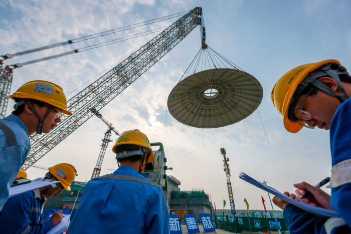 A group of workers wearing yellow safety helmets and blue work attire are seen making observations and taking notes at a construction site. Above them, a massive crane hoists a large circular component