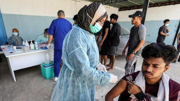 People line up at a vaccination centre, with a vaccine-giver injecting a young man in the foreground