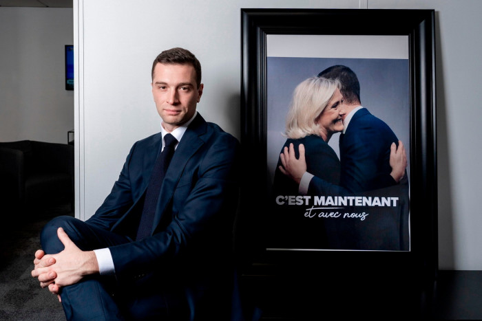 Jordan Bardella in front of a photograph showing him and Marine Le Pen hugging