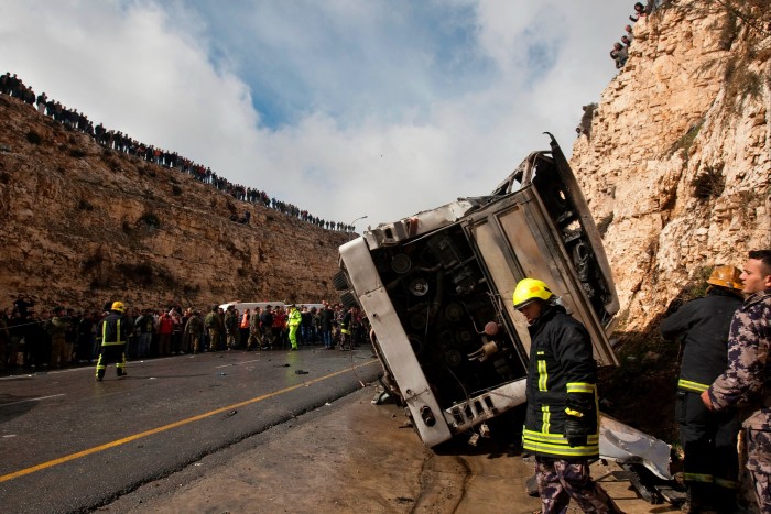 In a photo dated February 2012, an overturned bus lies on the side of a road in a ravine. Rescue workers in high-vis inspect the vehicle, while high on the top of the ravine stand dozens of onlookers