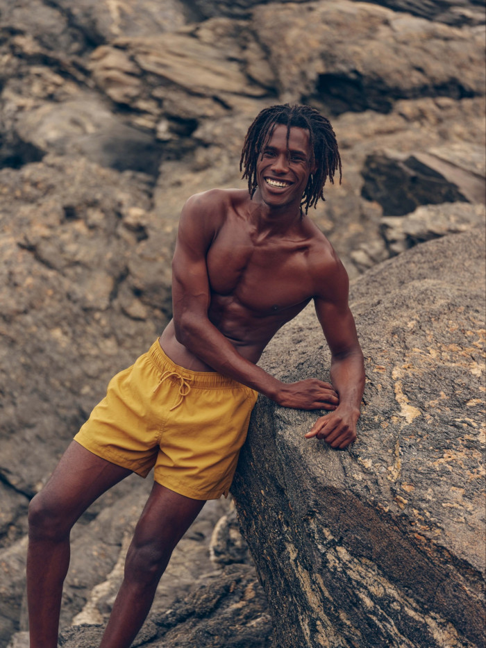 A young smiling man in yellow swim shorts leans against a rock