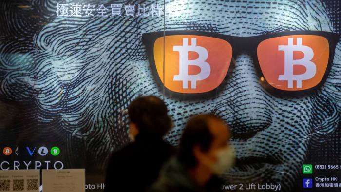 A display of cryptocurrency Bitcoin in Hong Kong. A big poster of a close up of image of George Washington from the dollar bill wearing darkglasses with the Bitcoin logo on the lenses. Two people are walking in front of the poster