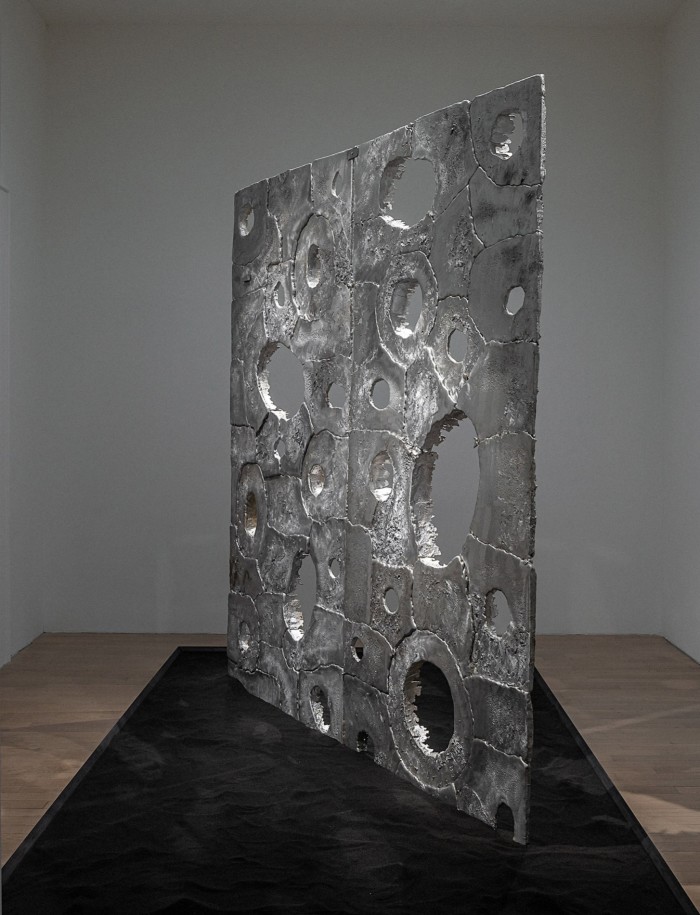 A metallic silver sculpture pierced with holes, installed in a gallery
