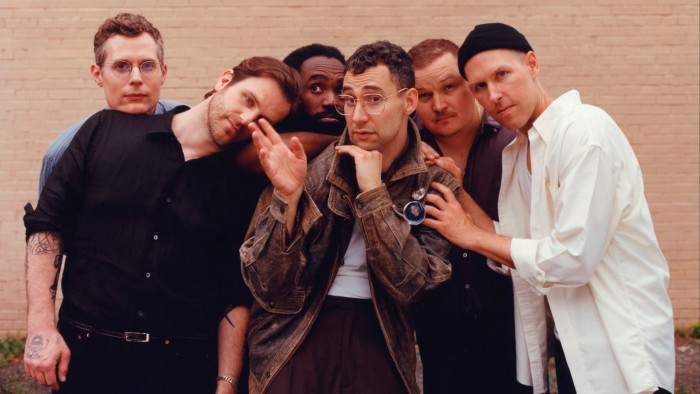 Five men huddle around Jack Antonoff, who poses with his hand under his chin, in front of a beige brick wall 
