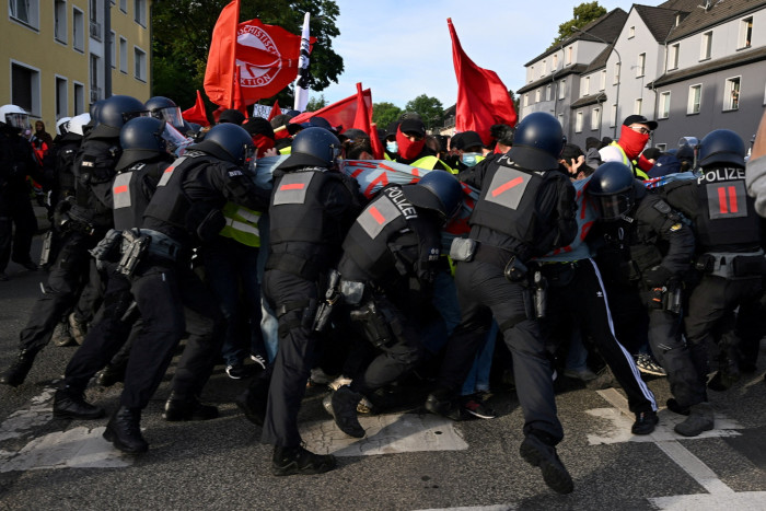 Figures dressed in black uniforms and black helmets, seen from behind, push against a crowd of people waving red flags