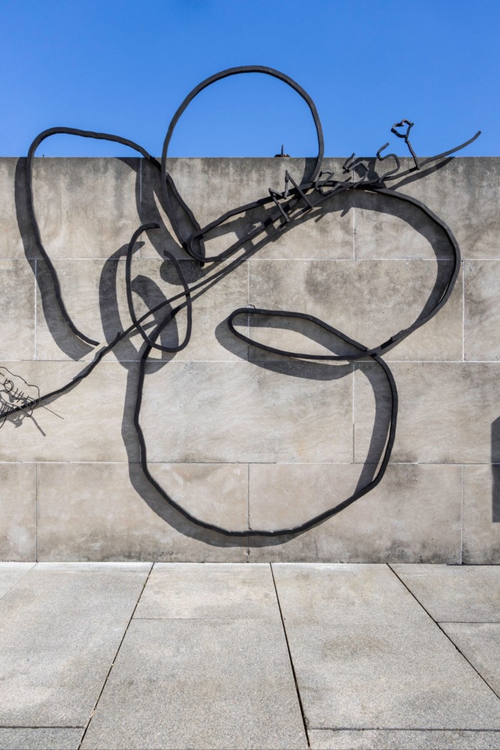 A metal sculpture in the shape of a simple flower is placed against a concrete wall